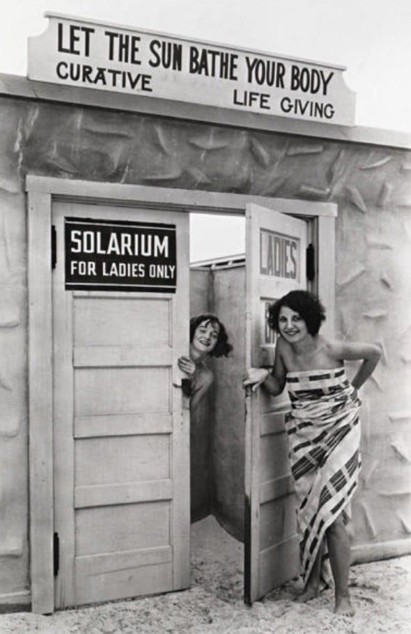 Two women in towels stand at the entrance of a “Ladies Only” solarium. Above the door, a sign reads, “Let the sun bathe your body – curative, life giving.” The scene appears vintage, in black and white.