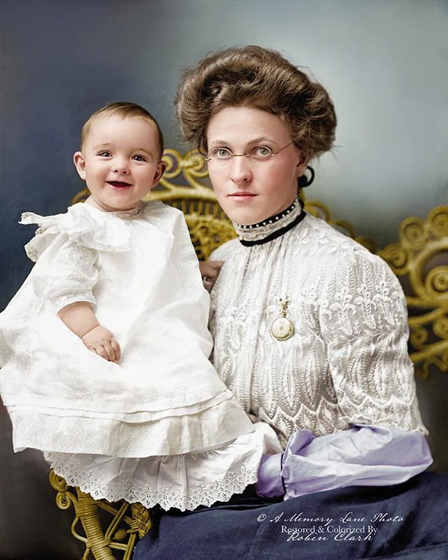 A woman in Victorian-era clothing and glasses sits on a yellow ornate chair, holding a smiling baby dressed in a white gown. The photo is colorized and has a soft blue and gray background.
