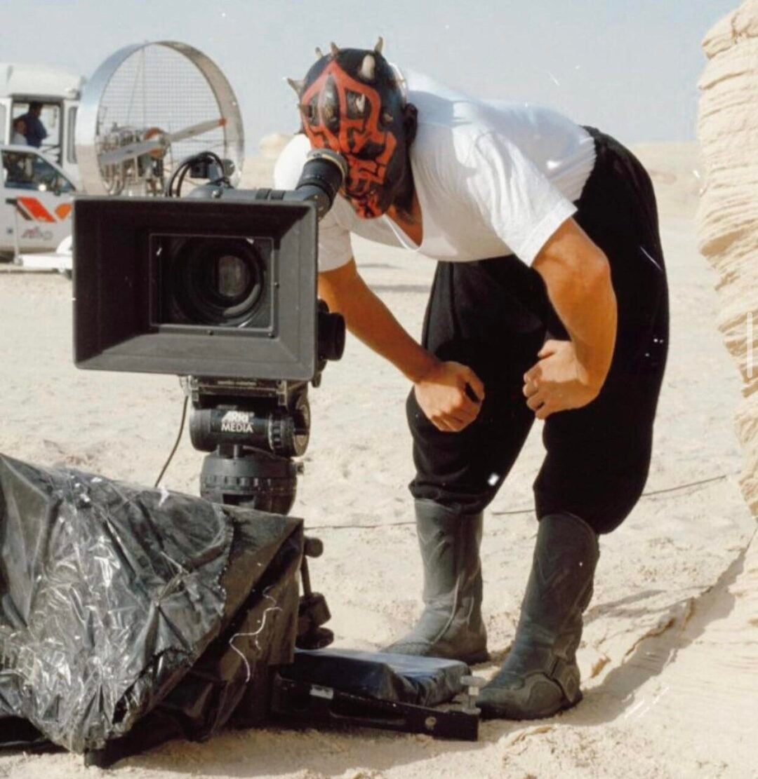 A person wearing a Darth Maul mask looks through a film camera on a sandy outdoor set, dressed in casual clothes and boots. Film equipment and a vehicle are visible in the background.