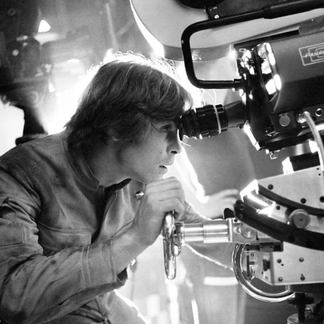 A young man in a costume looks through the viewfinder of a large film camera on a movie set, illuminated by bright studio lights. The image is in black and white.