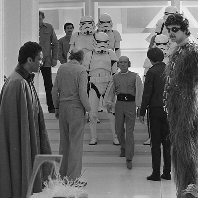 A black-and-white behind-the-scenes photo showing actors in Star Wars costumes, including Stormtroopers, Chewbacca, and Lando Calrissian, along with crew members on a sci-fi film set.