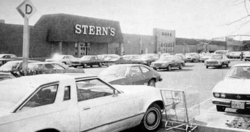 Black-and-white photo of a parking lot filled with cars in front of a Stern’s department store. A shopping cart is near the front, and the store's sign is prominently visible on the building.