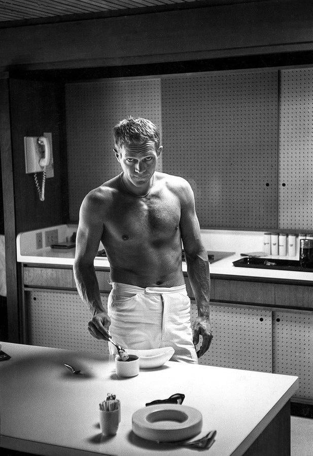 A shirtless man in white pants stands in a modern kitchen, holding a spoon over a bowl on a counter. The scene is in black and white, and the man looks down with a focused expression.