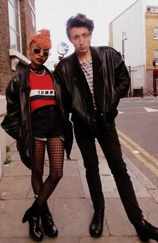 Two people stand on a city sidewalk wearing black leather jackets. One has orange hair, sunglasses, a red Tommy Jeans shirt, fishnet tights, and boots. The other has short dark hair, a striped shirt, black pants, and boots.