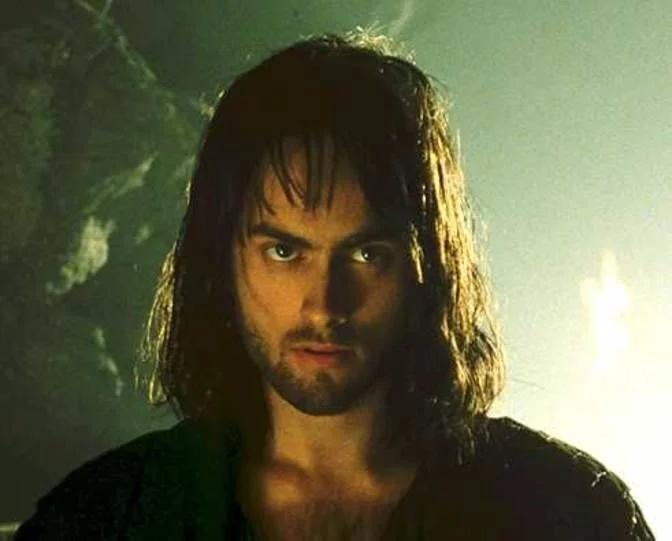 A person with long, dark hair and a beard looks intensely at the camera. The background is dimly lit with a soft, greenish hue, creating a mysterious atmosphere.