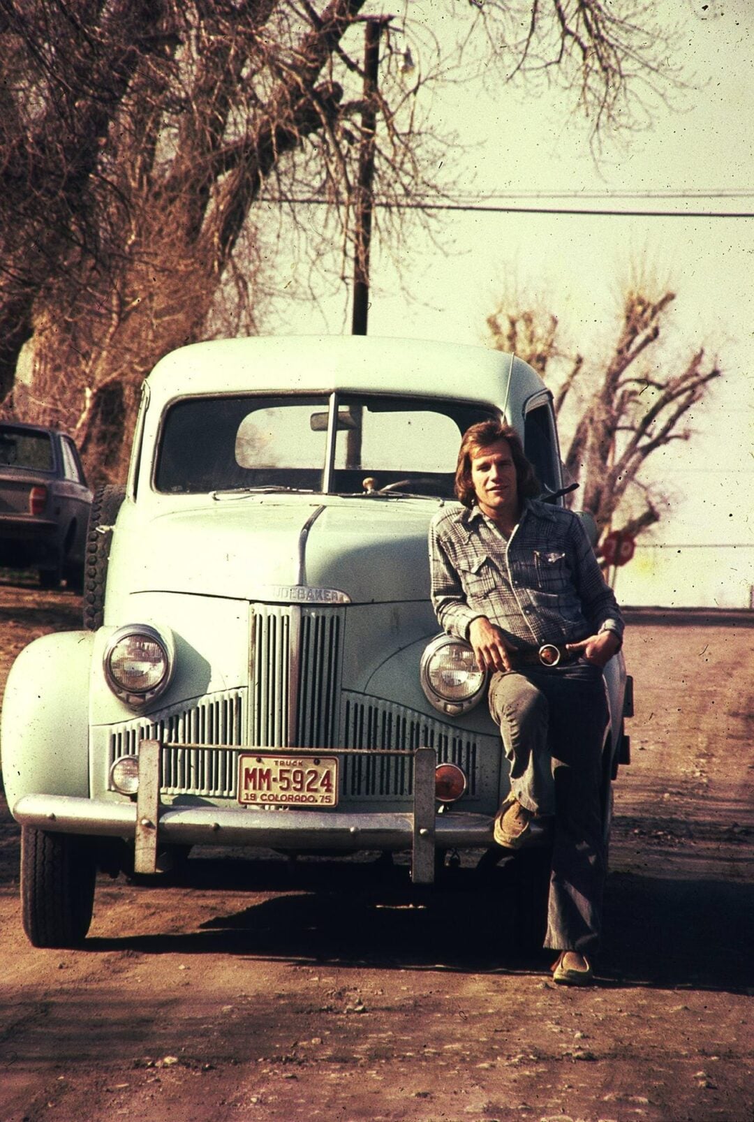 A man in a plaid shirt and jeans leans against the front of a vintage light blue car parked on a quiet, tree-lined street with bare branches, under an overcast sky.