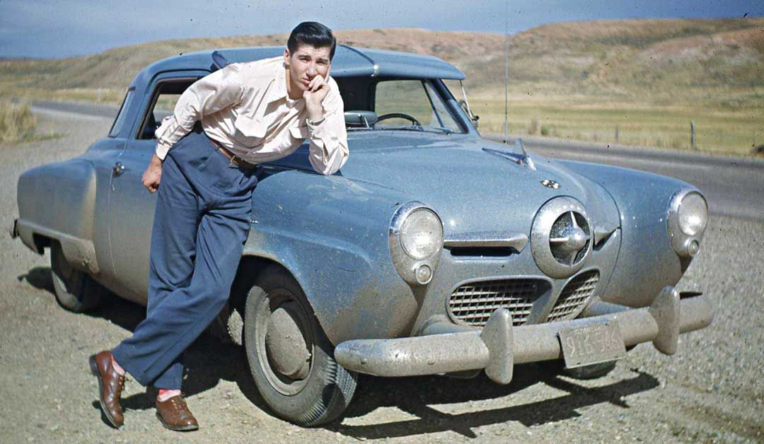 A man in a light shirt and dark pants leans on the front of a vintage blue car parked on a gravel road, with dry hills and open landscape in the background.