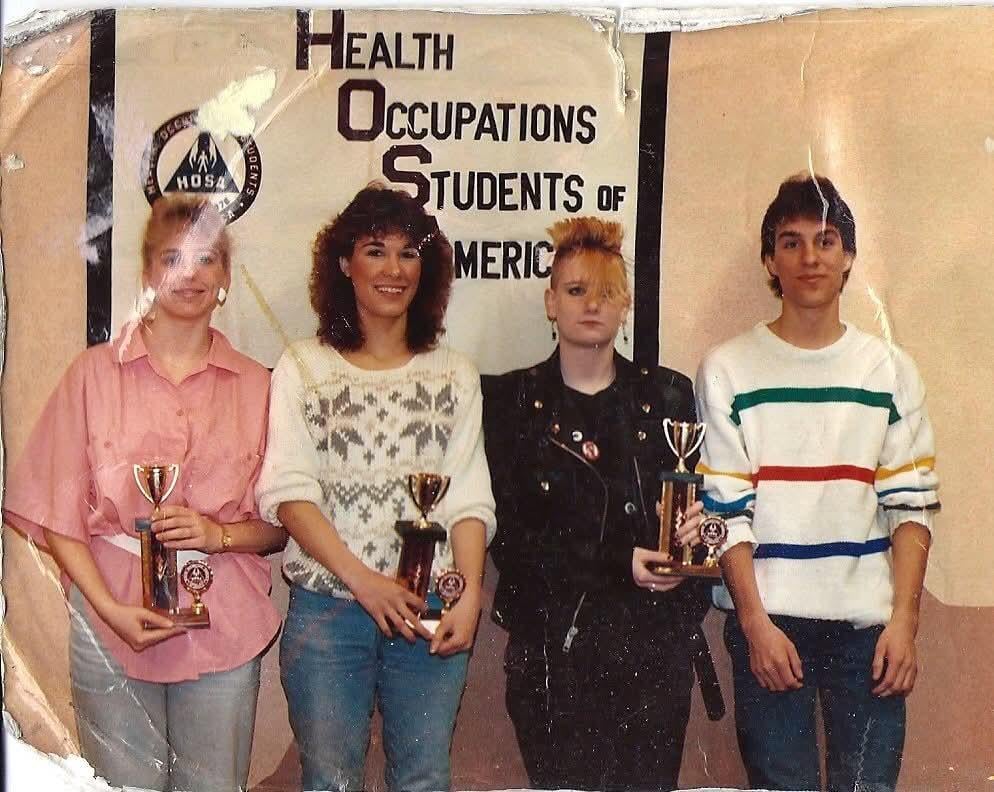Four young people stand in front of a "Health Occupations Students of America" sign, each holding a trophy. They are dressed in 1980s-style clothing and smiling, except one person in a leather jacket with a serious expression.