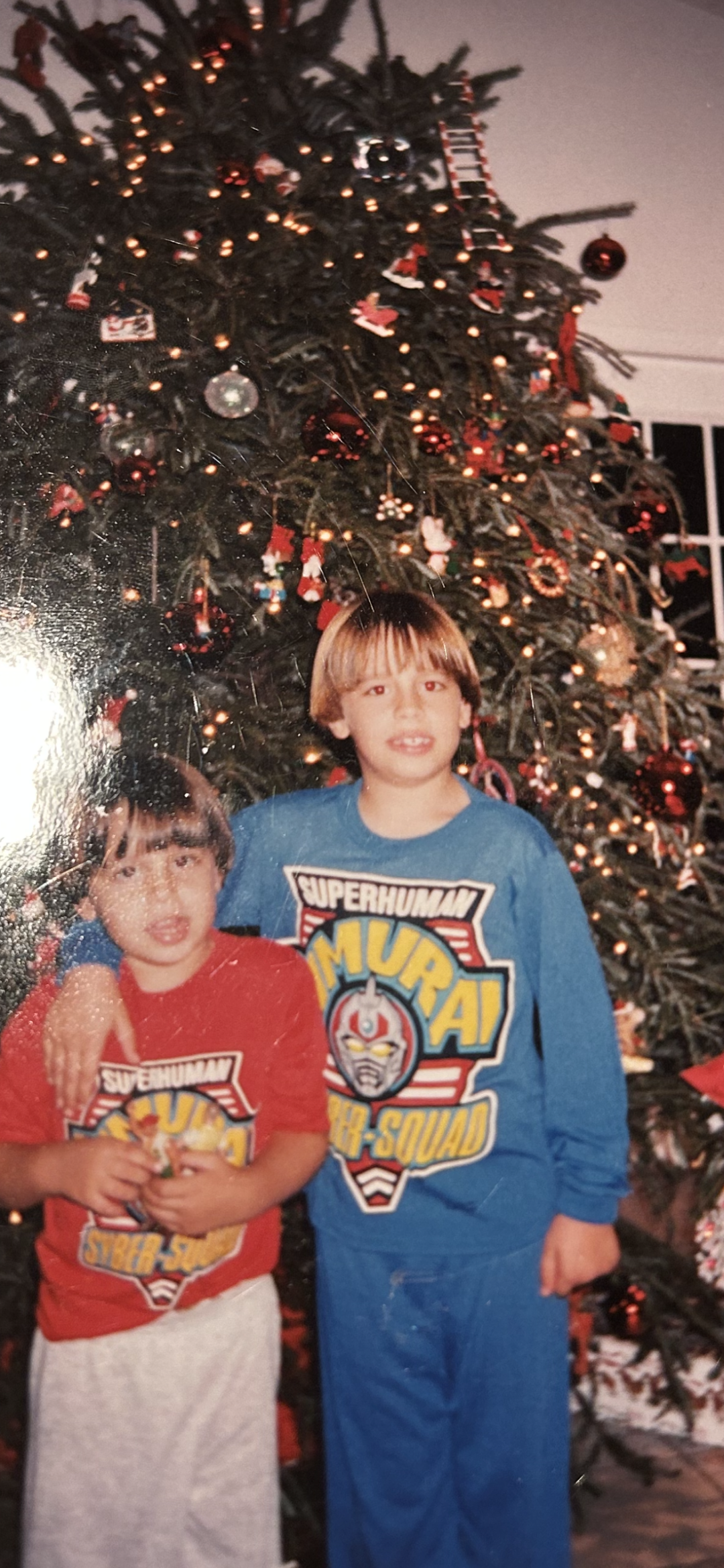 Two children wearing matching Superhuman Samurai Syber-Squad pajamas stand in front of a decorated Christmas tree. One child has their arm around the other, and both are smiling.