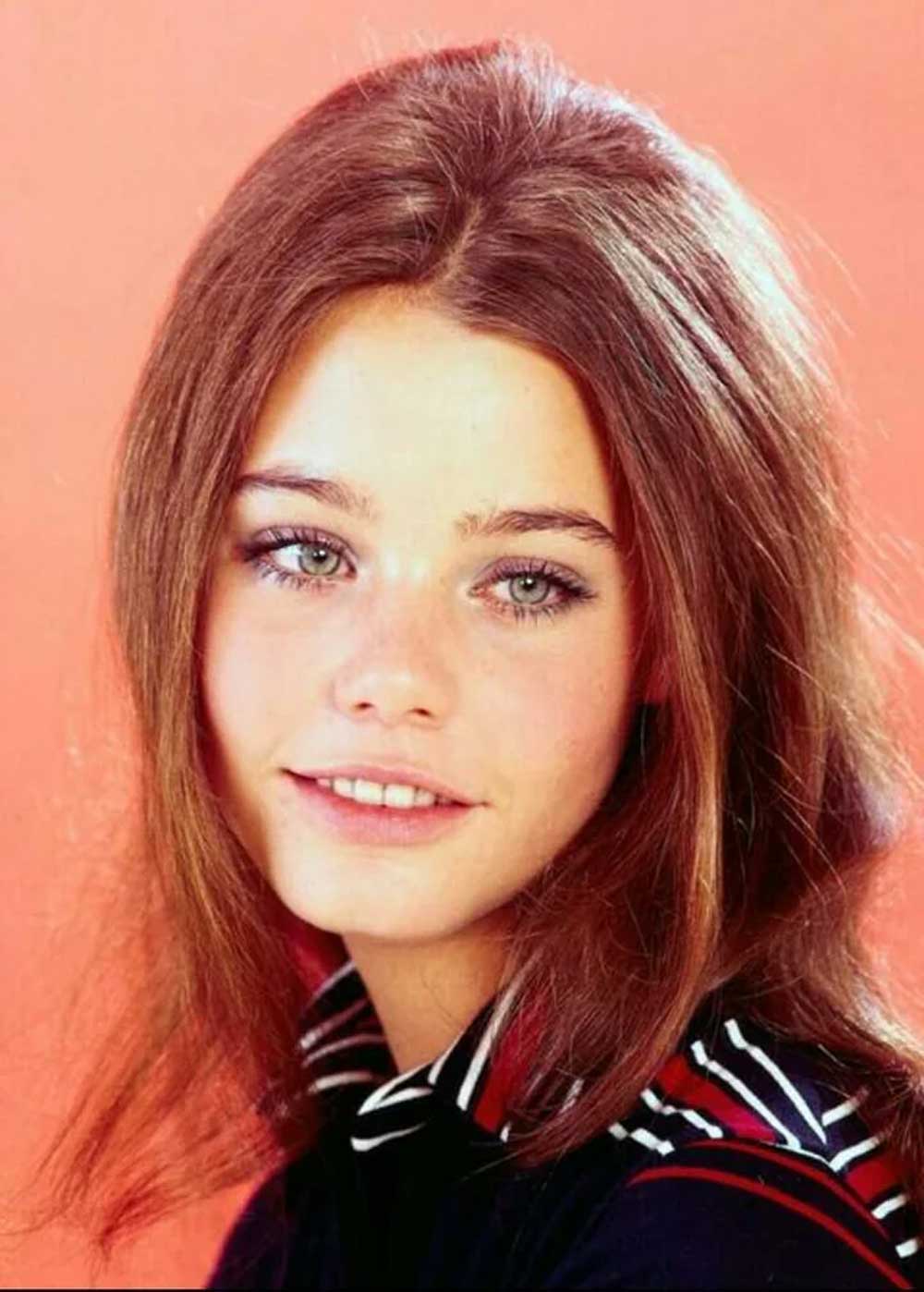 A young woman with straight, light brown hair and light-colored eyes poses in front of a peach background. She is wearing a black top with red and white stripes and is smiling softly at the camera.