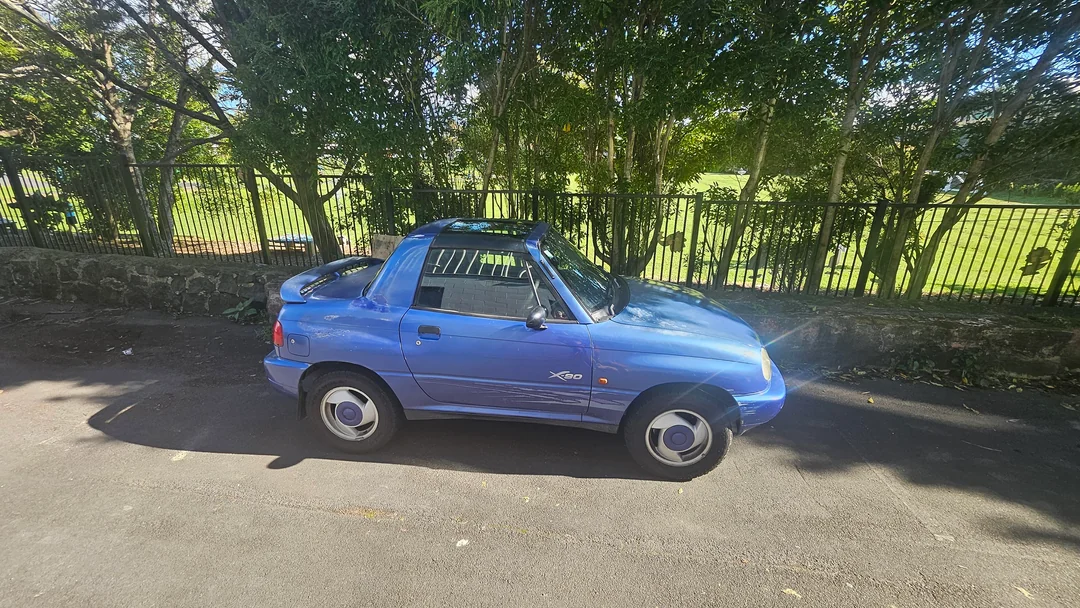 A blue Suzuki X-90 compact car is parked on a shaded street near a black metal fence and green trees on a sunny day.