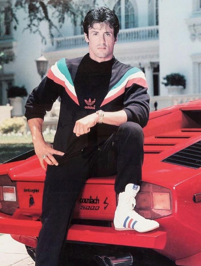 A man in a black Adidas tracksuit and white sneakers poses with one foot on a red Lamborghini Countach 5000 S, in front of a large white mansion.