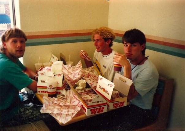 Three young men sit at a Taco Bell booth with a table full of food wrappers, cups, and boxes, appearing to enjoy a large fast food meal together. The setting looks casual and retro.