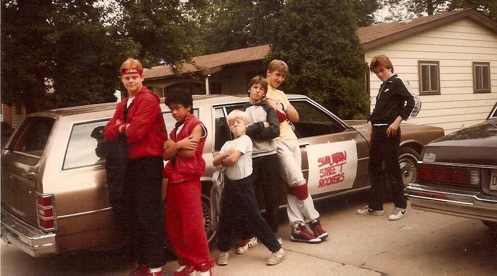 Six kids pose confidently in front of a brown station wagon labeled "Sugar Shock Rockers," with houses and trees in the background. They wear 1980s clothing, some with headbands and jackets, standing with arms crossed or hands on hips.