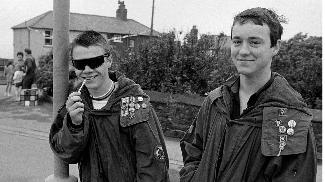 Two teenage boys wearing jackets with multiple pins and badges stand on a sidewalk. One boy wears dark sunglasses and holds a pen to his mouth, while the other smiles. Houses and bushes are visible in the background.