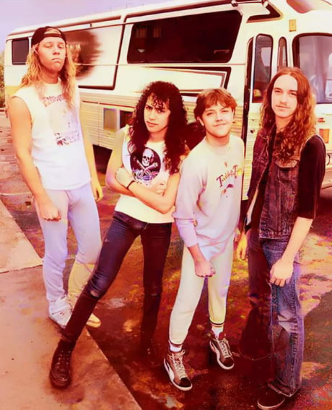 Four young men with long hair pose in front of a tour bus. Three wear casual clothes, including jeans and t-shirts, while one wears a sleeveless vest and jeans. All stand on pavement, looking at the camera.