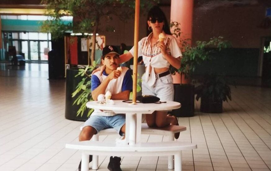 Two people eating ice cream at a white picnic table inside a mall. One is sitting and wearing a baseball cap and shorts; the other is standing behind, wearing sunglasses, a tied shirt, and shorts. There are plants and columns in the background.