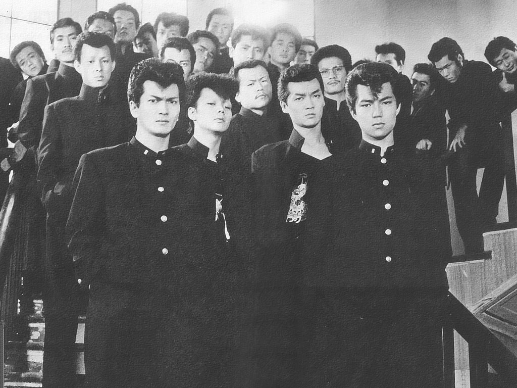A group of serious-looking young men in matching dark school uniforms stand and sit on a staircase, posing closely together for a black-and-white photograph.