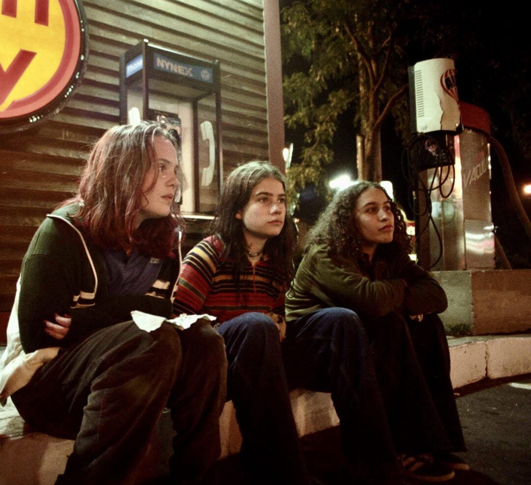 Three young people sit on a curb at night near a payphone and a gas station, looking thoughtful. Dim lights illuminate the scene, creating a nostalgic, moody atmosphere.