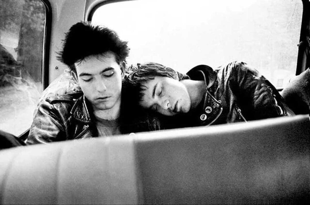 Two young men in leather jackets sit in the backseat of a car. One is resting his head on the other's shoulder, both appearing tired or relaxed. The image is black and white.