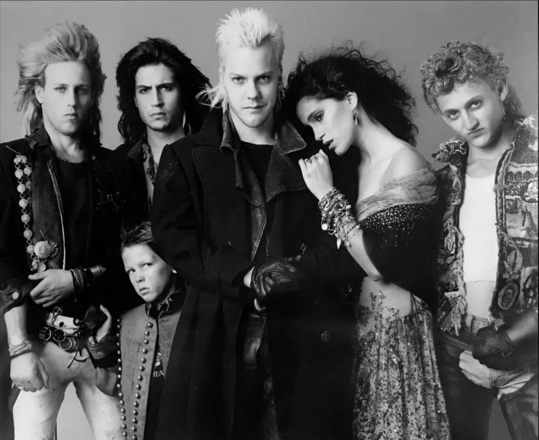 A black-and-white photo of five men and one woman with 1980s punk and goth styles, featuring dramatic hairstyles, makeup, and layered clothing, posing closely together and looking at the camera.