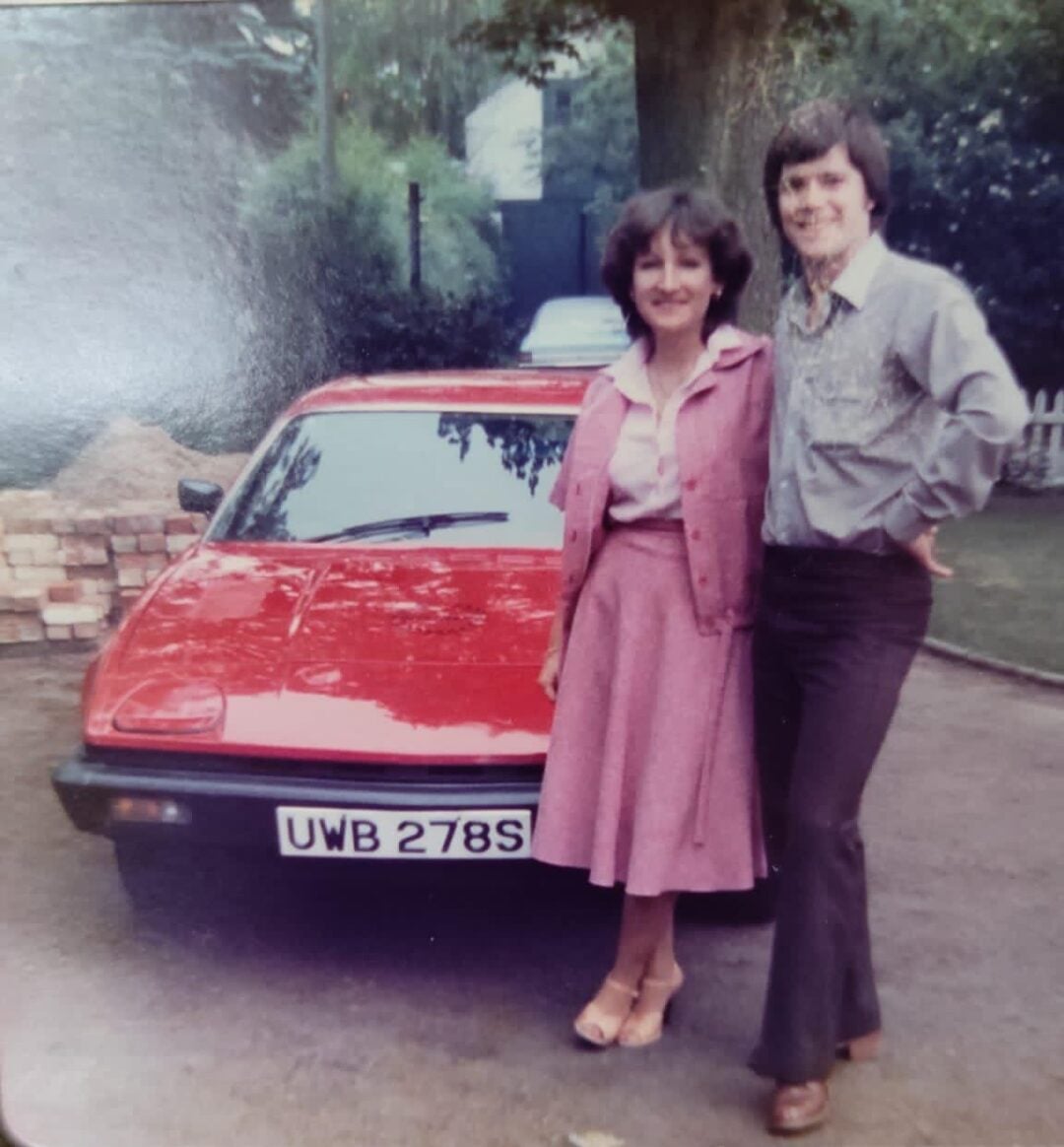 A smiling woman in a pink skirt suit and a man in a patterned shirt and brown pants stand in front of a red vintage car with license plate UWB 278S, outside on a driveway with greenery in the background.