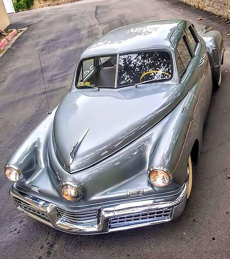 A silver vintage car with a unique, rounded design and prominent front headlights is parked on a paved street. The car has a classic look with a sleek, smooth body and chrome details.