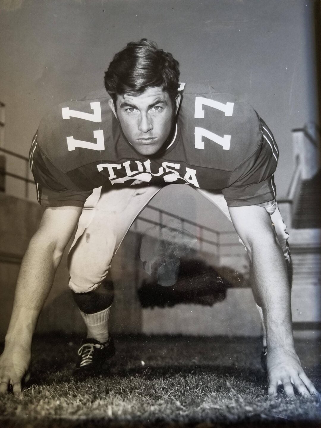 Black and white photo of a football player in a three-point stance, wearing a Tulsa jersey with the number 77. The athlete is looking forward with a focused expression, posed outdoors near stadium seating.