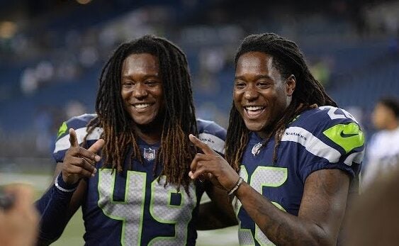 Two football players in Seattle Seahawks uniforms, numbers 49 and 26, smile and pose together on a football field, holding up their index fingers. The stadium seats are blurred in the background.