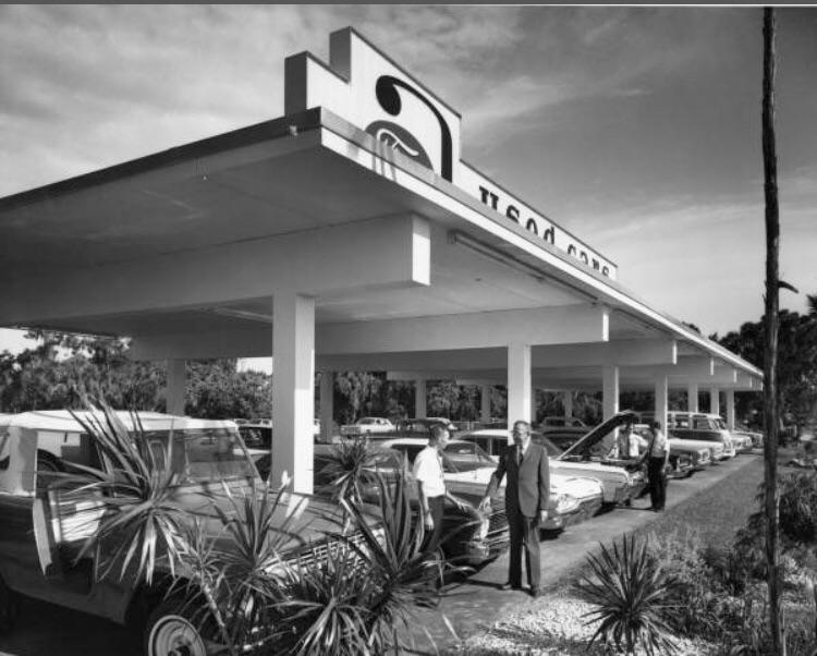 Black-and-white photo of a mid-century carport with several vintage cars parked underneath. Two men stand near the vehicles, and palm plants are visible in the foreground. A sign with stylized text is on the roof.