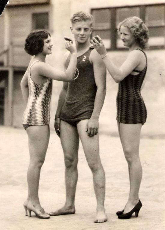 Three young adults in vintage swimsuits pose playfully; two women, one on each side, touch the chin and cheek of a smiling man standing between them. Both women wear heels. Outdoor setting, likely from the early 20th century.