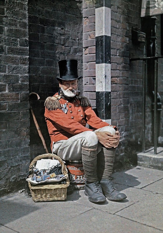 An elderly man with a white beard wearing a red uniform with gold epaulets and a black top hat sits on a step beside a brick wall, with a cane and a basket of assorted items at his side.