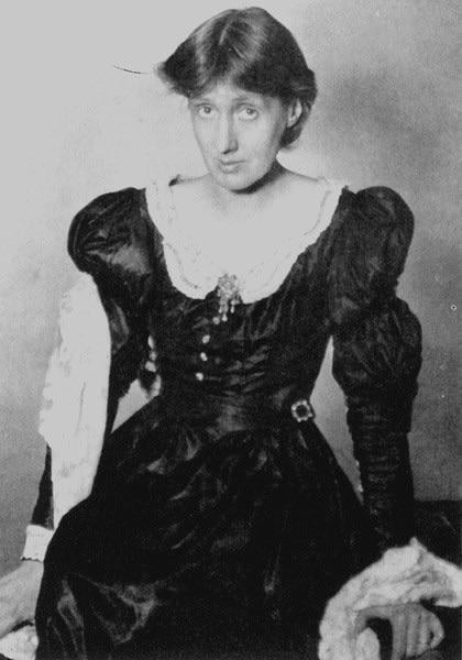 A black-and-white portrait of a woman with short hair, wearing a dark, long-sleeved, Victorian-style dress with puffed shoulders and lace details, sitting and looking slightly upward.