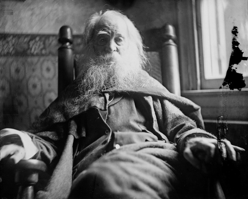 An elderly man with a long white beard sits in a wooden rocking chair, wearing a thick coat. The black-and-white photo has a vintage feel, with light streaming through a window behind him.