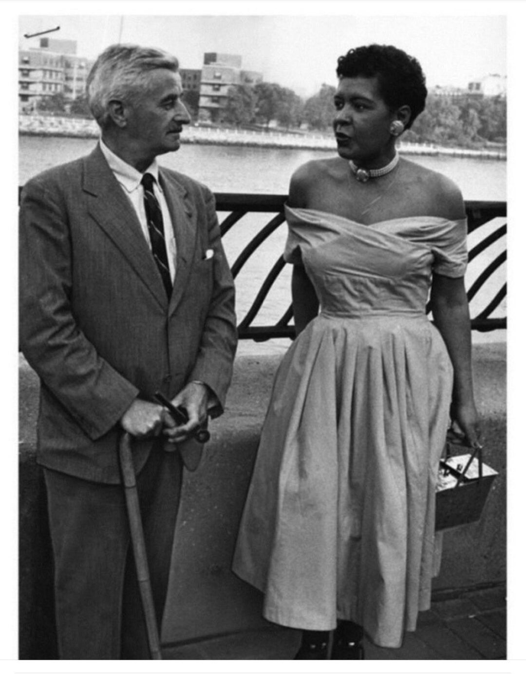 A man in a suit holding a cane stands beside a woman in an off-the-shoulder dress and necklace. They are talking outdoors near a railing with water and buildings in the background.
