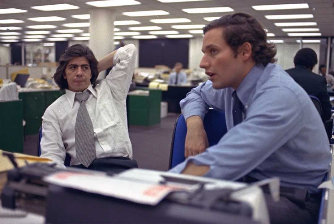 Two men in dress shirts and ties sit in a busy office with fluorescent lighting. One leans back with his hand behind his head, while the other sits forward, engaged in conversation. Office desks and papers are visible in the background.