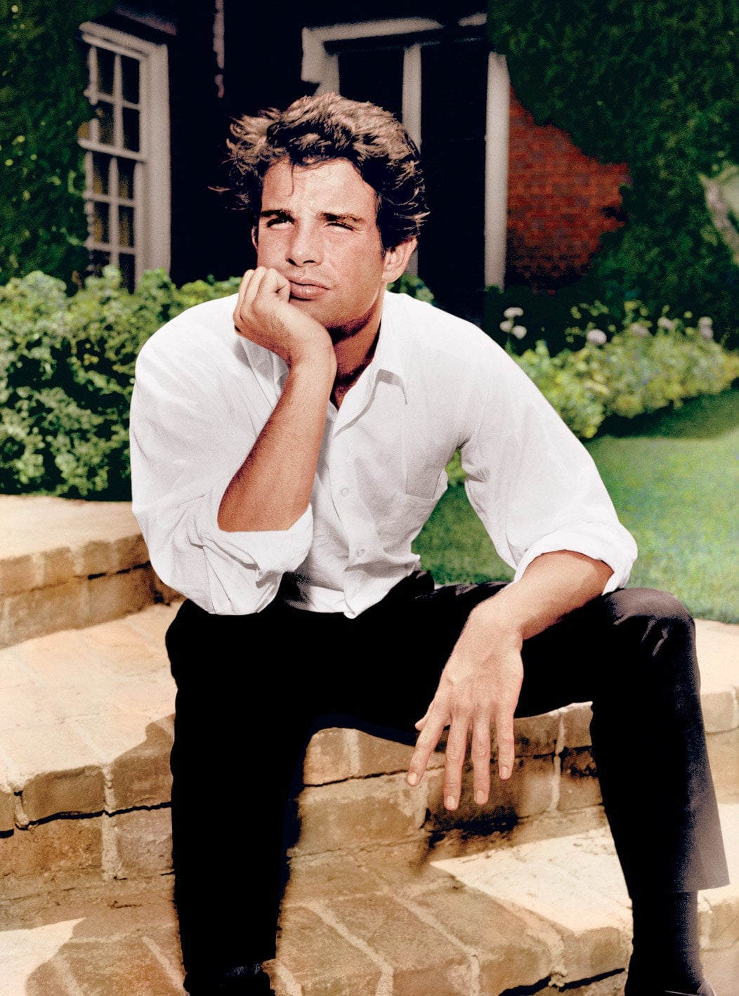 A man in a white shirt and dark pants sits on brick steps outside, resting his chin on his hand and looking thoughtfully into the distance. Green grass and a house are visible in the background.