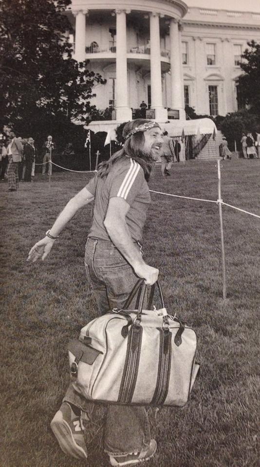 A man wearing a headband, t-shirt, jeans, and sneakers carries a duffel bag while walking on the White House lawn, looking back and smiling. The White House columns are visible in the background.