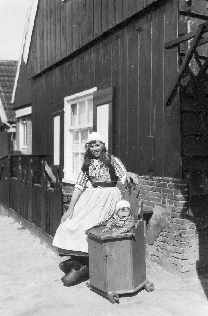 A woman in traditional Dutch clothing and wooden shoes stands next to a small child seated in a wooden cart outside a dark wooden house with brick foundation. Both are smiling, and the setting appears historic or early 20th century.