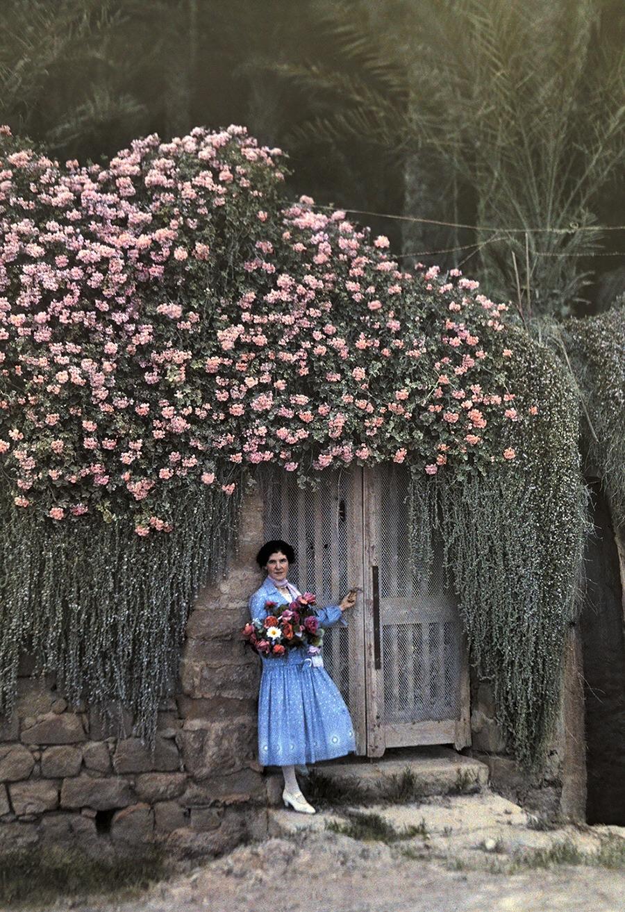 A woman in a blue dress holding a bouquet stands in front of a rustic wooden door framed by a stone wall and covered with abundant pink flowering vines.