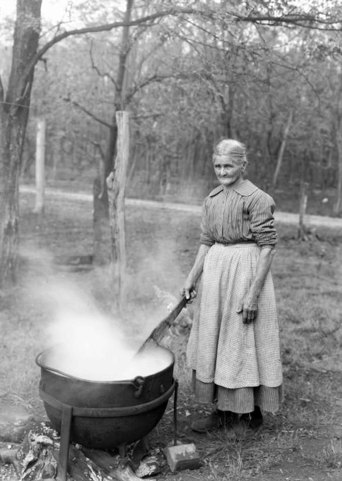 An older woman in a long dress and apron stands outdoors stirring a steaming cauldron over a fire, surrounded by trees and a rustic wooden fence in the background.