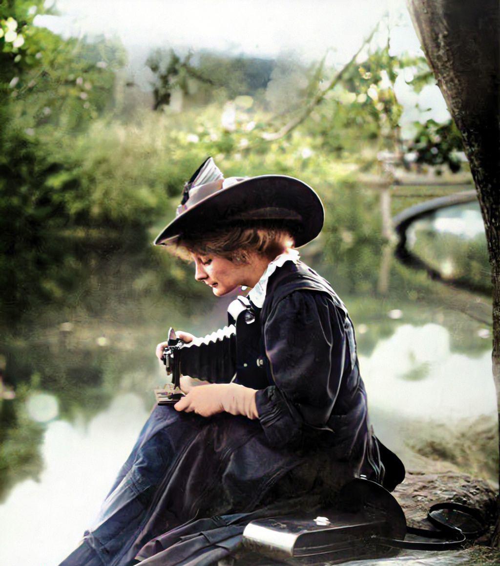 A woman in vintage clothing and a wide-brimmed hat sits by a calm riverside, looking down and adjusting an old-fashioned box camera on her lap. Trees and a footbridge are visible in the background.