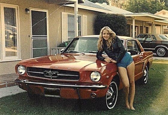 A young woman with long blonde hair, wearing a black jacket and blue shorts, leans against a red classic Ford Mustang parked on a grassy lawn in front of a suburban house.