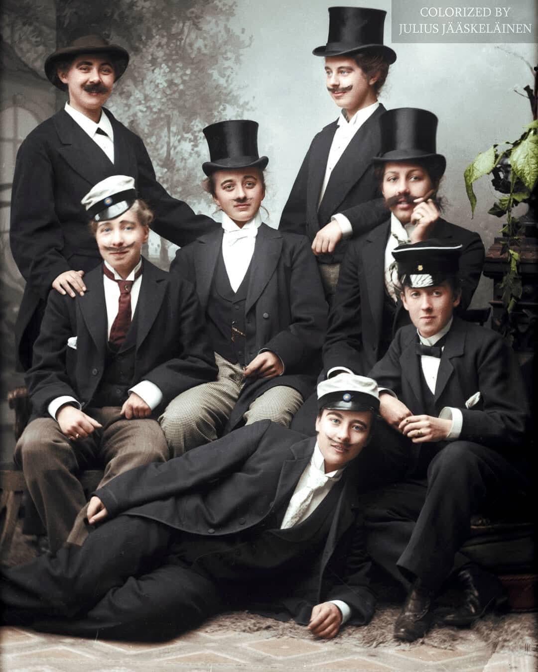 Seven young women dressed in suits and hats pose for a formal group portrait, some seated and others standing, with neutral expressions against a studio backdrop. The image is colorized and marked by Julius Jääskeläinen.