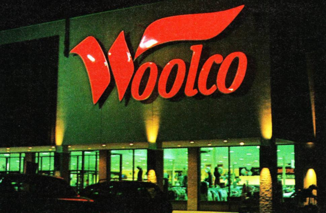 A brightly lit Woolco store at night, with a large red Woolco sign above the entrance and display windows showing clothing and other merchandise inside.
