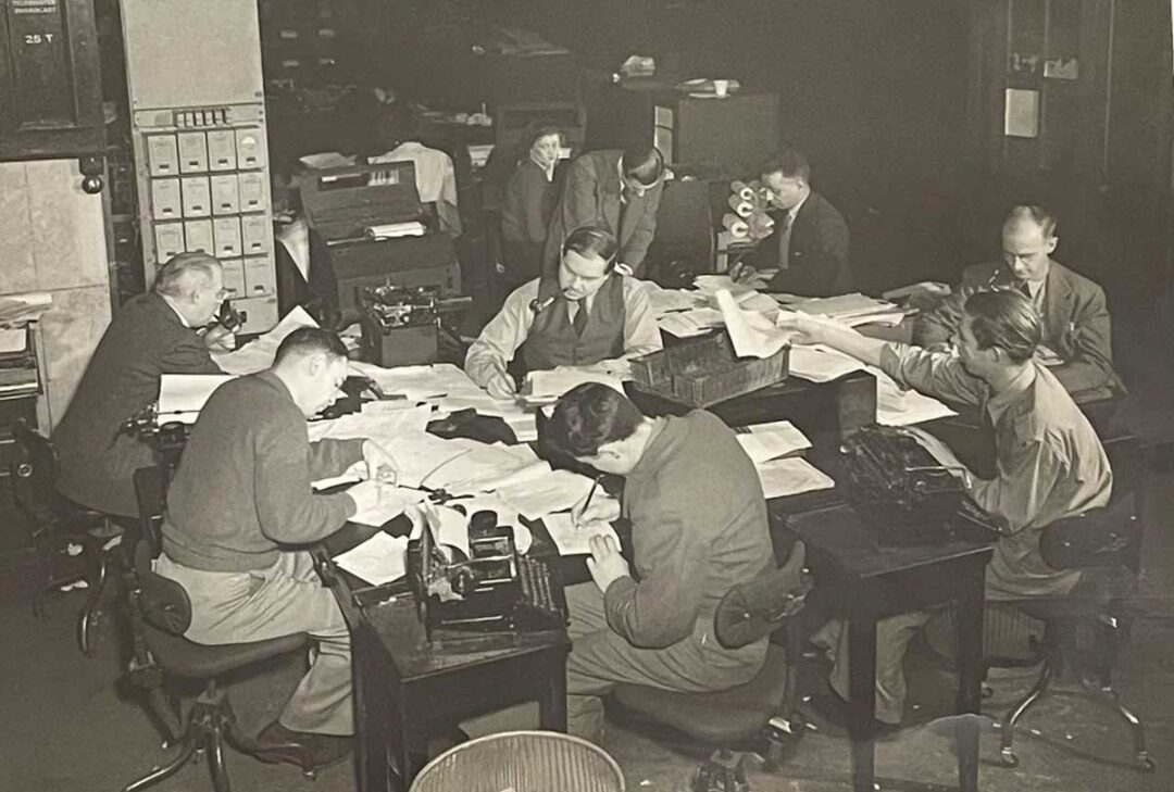A group of men and women work at desks covered with papers and documents in a busy, old-fashioned office setting, possibly from the mid-20th century. Typewriters and filing cabinets are visible in the background.