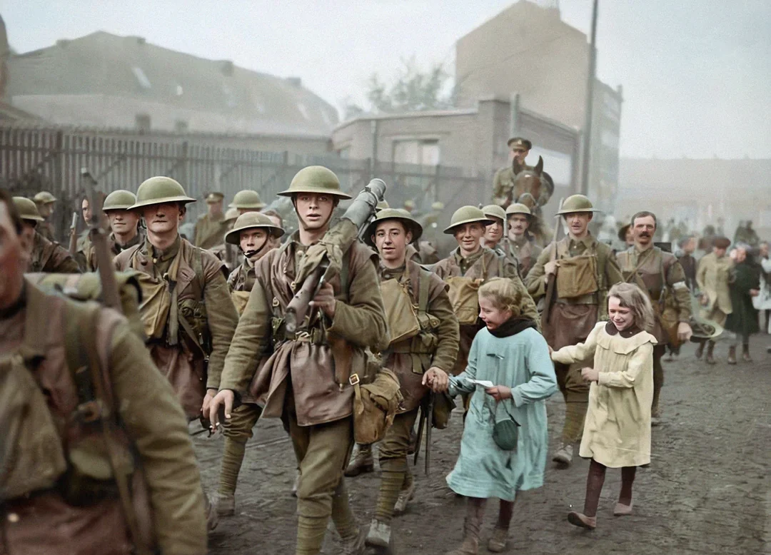 A group of World War II soldiers in uniform march down a cobblestone street, some smiling; two young girls in dresses walk alongside, holding hands with one of the soldiers.