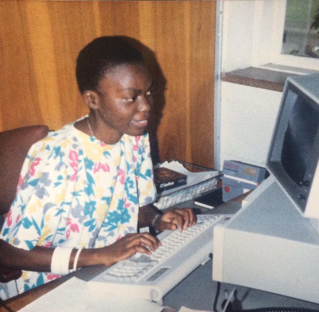 A woman in a floral dress types on a vintage computer keyboard at a desk. She is seated indoors near a window, with office supplies and papers around her.