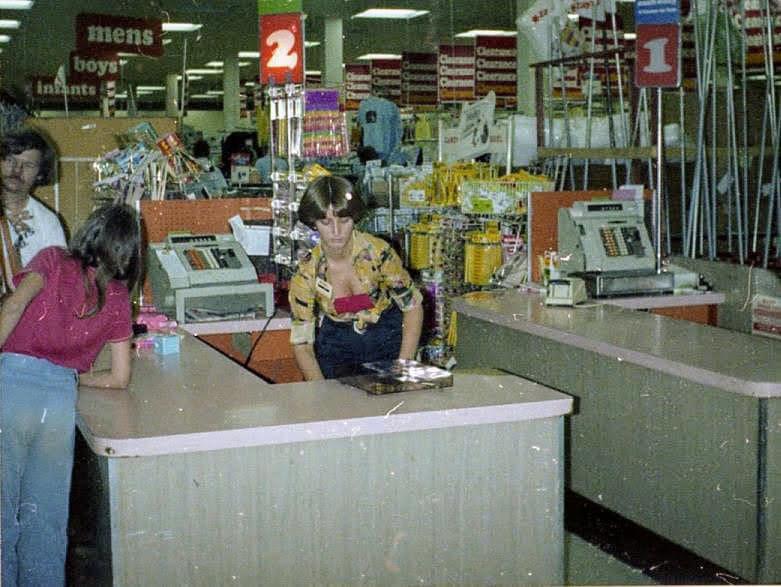 A vintage photo of a grocery store checkout area with two register lanes, cashiers, and customers. The area is busy and displays signs for mens, boys, infants, and clearance items in the background.