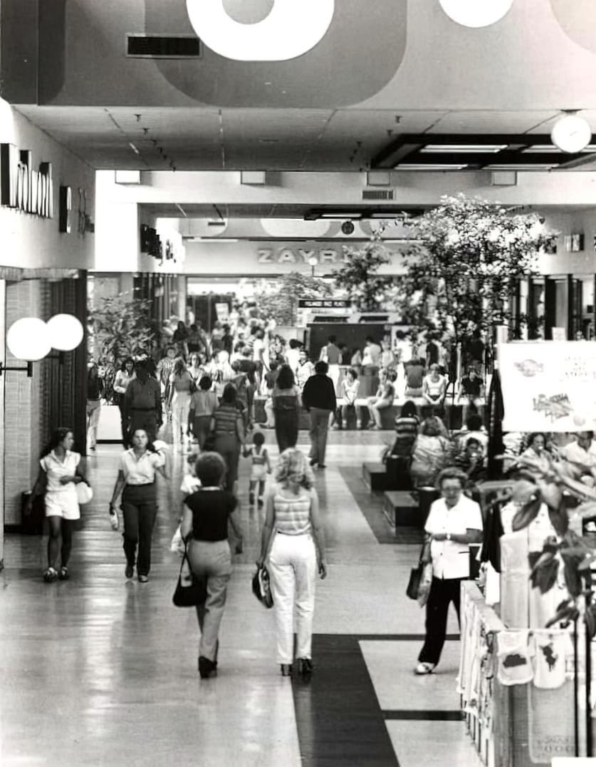 Black and white photo of a busy shopping mall in the 1970s, showing many people walking, shopping, and socializing. Stores line both sides, and a large “Zayre” sign is visible in the background.